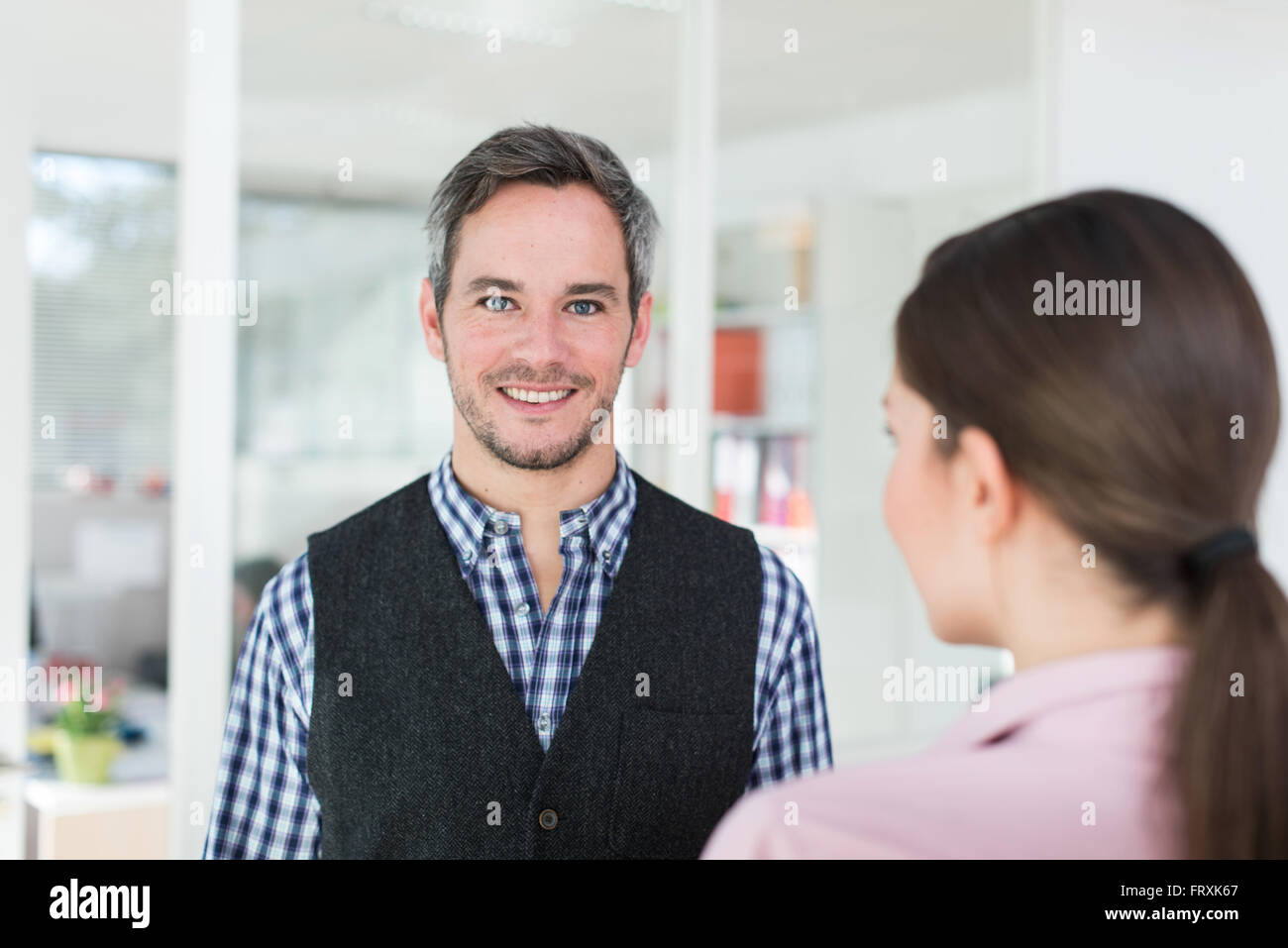 Portrait of a forty years old man with grey hair and beard wearing a checkered blue shirt. A woman with a pink shirt is facing him, focus on the man. They are standing in a luminous open space. Stock Photo