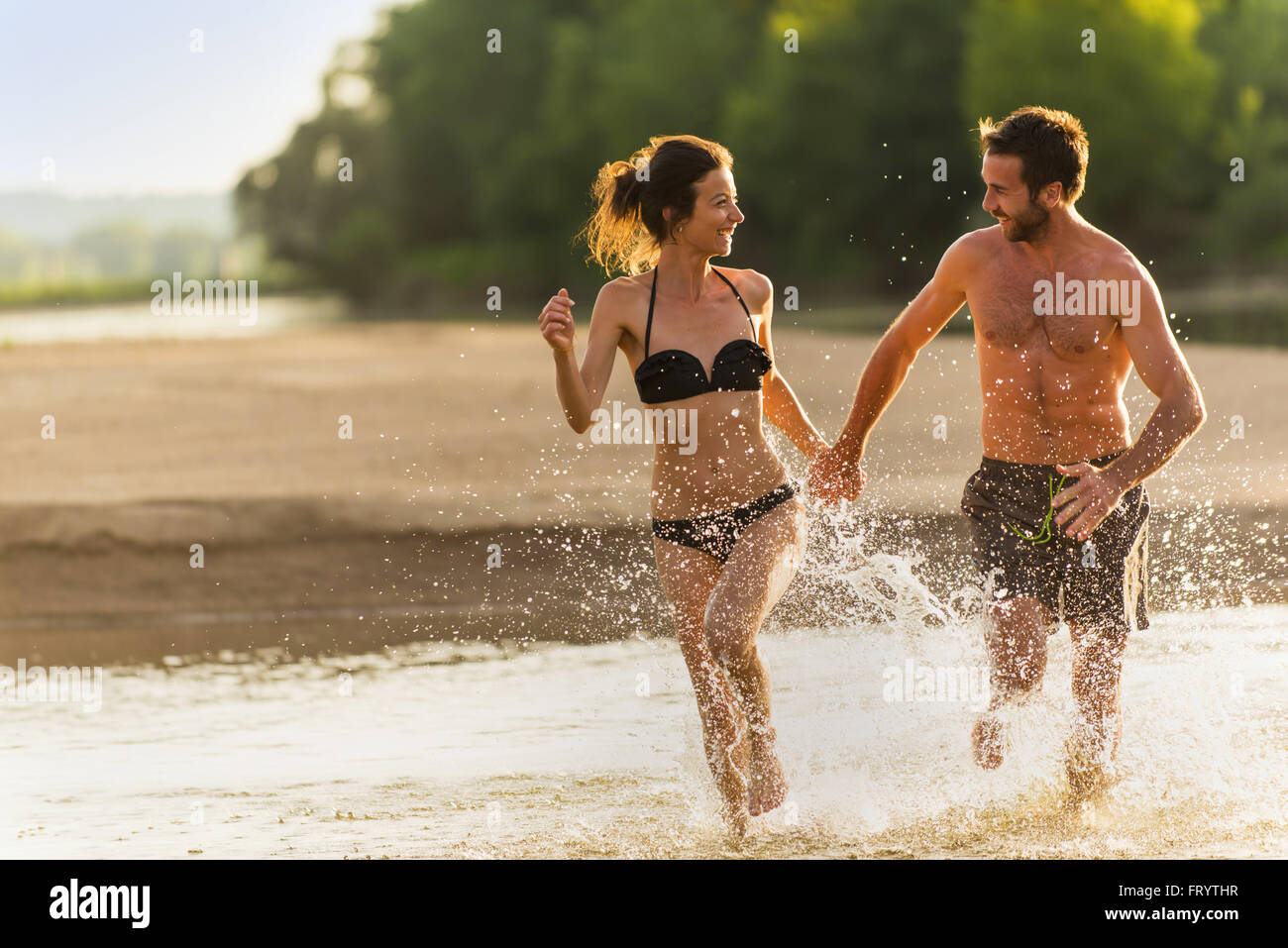 A nice couple in his 30's is holding hands and running in the water at the  beach during the holidays. The man is shirtless and the woman has a bikini.  The water