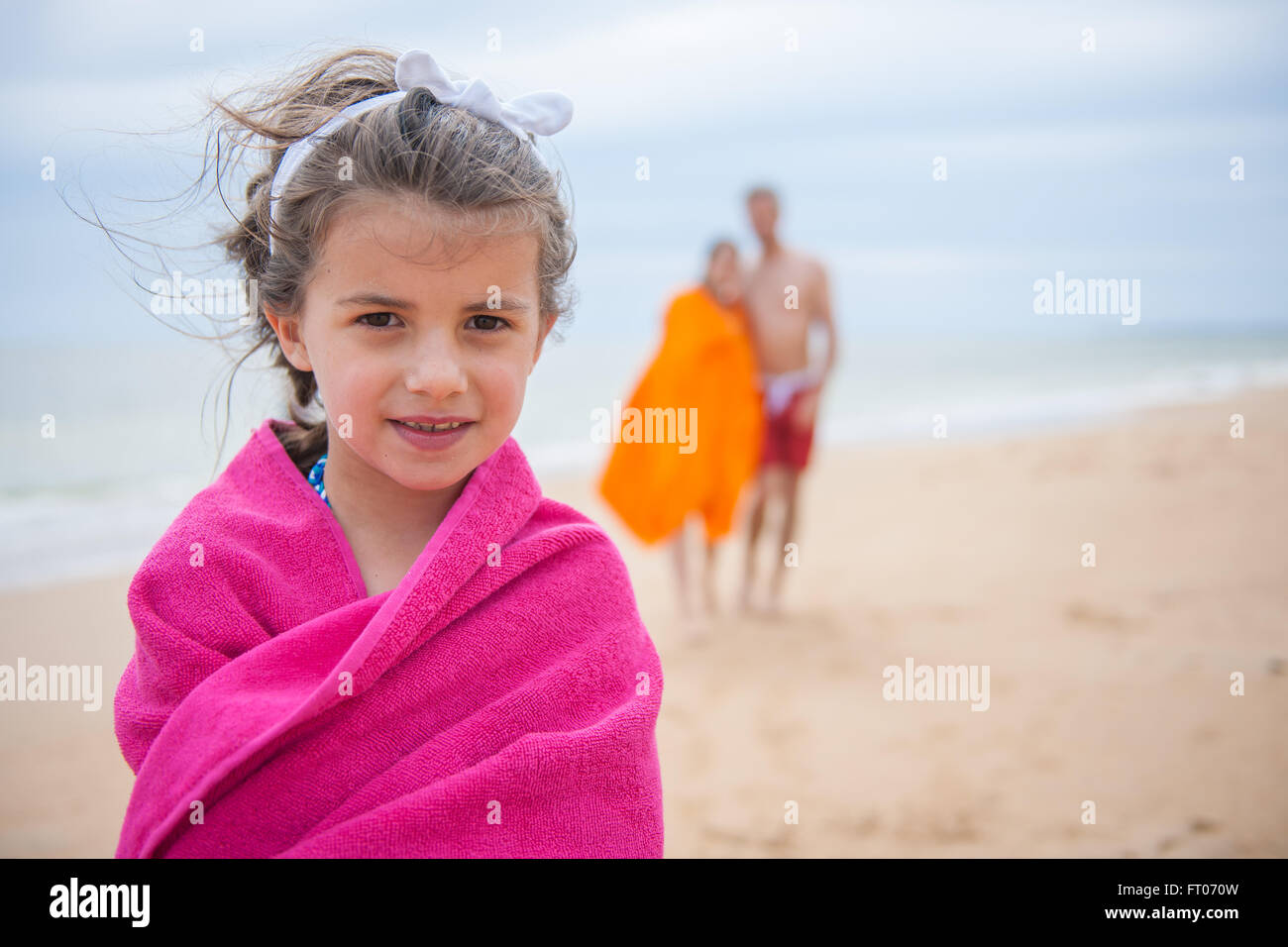Portrait of a six year old at the beach. She is wrapped in a pink towel, her parents are in the