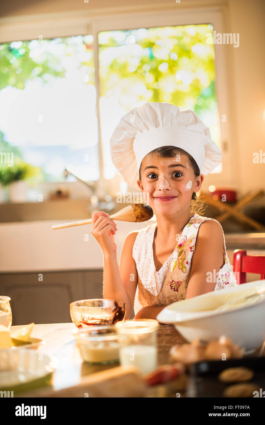 A seven years old girl with chef hat is holding a wooden spoon full of chocolate. She is looking at camera, sitting at a wooden Stock Photo