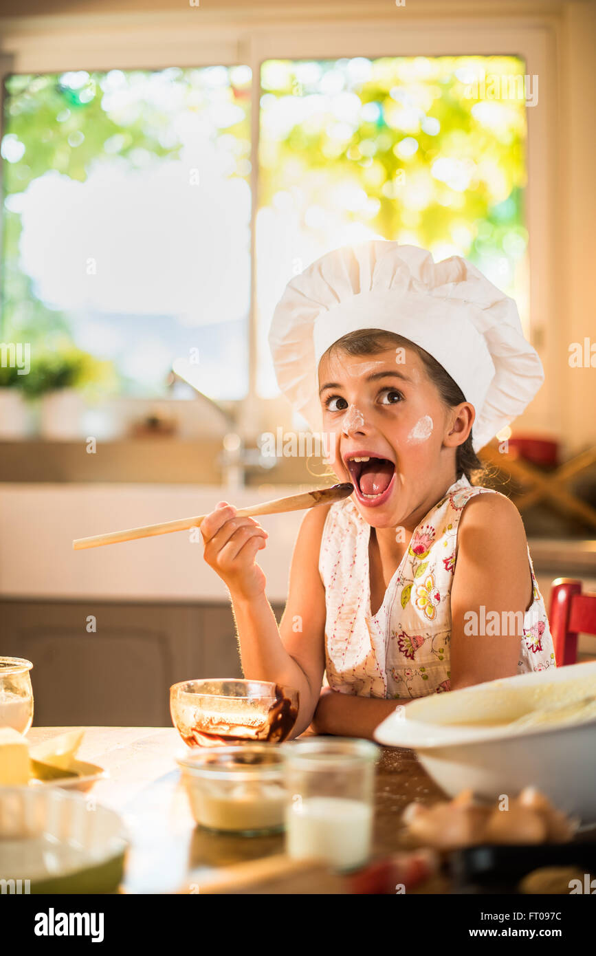 A seven years old girl with chef hat is licking a wooden spoon full of chocolate. She is looking at camera, sitting at a wooden Stock Photo