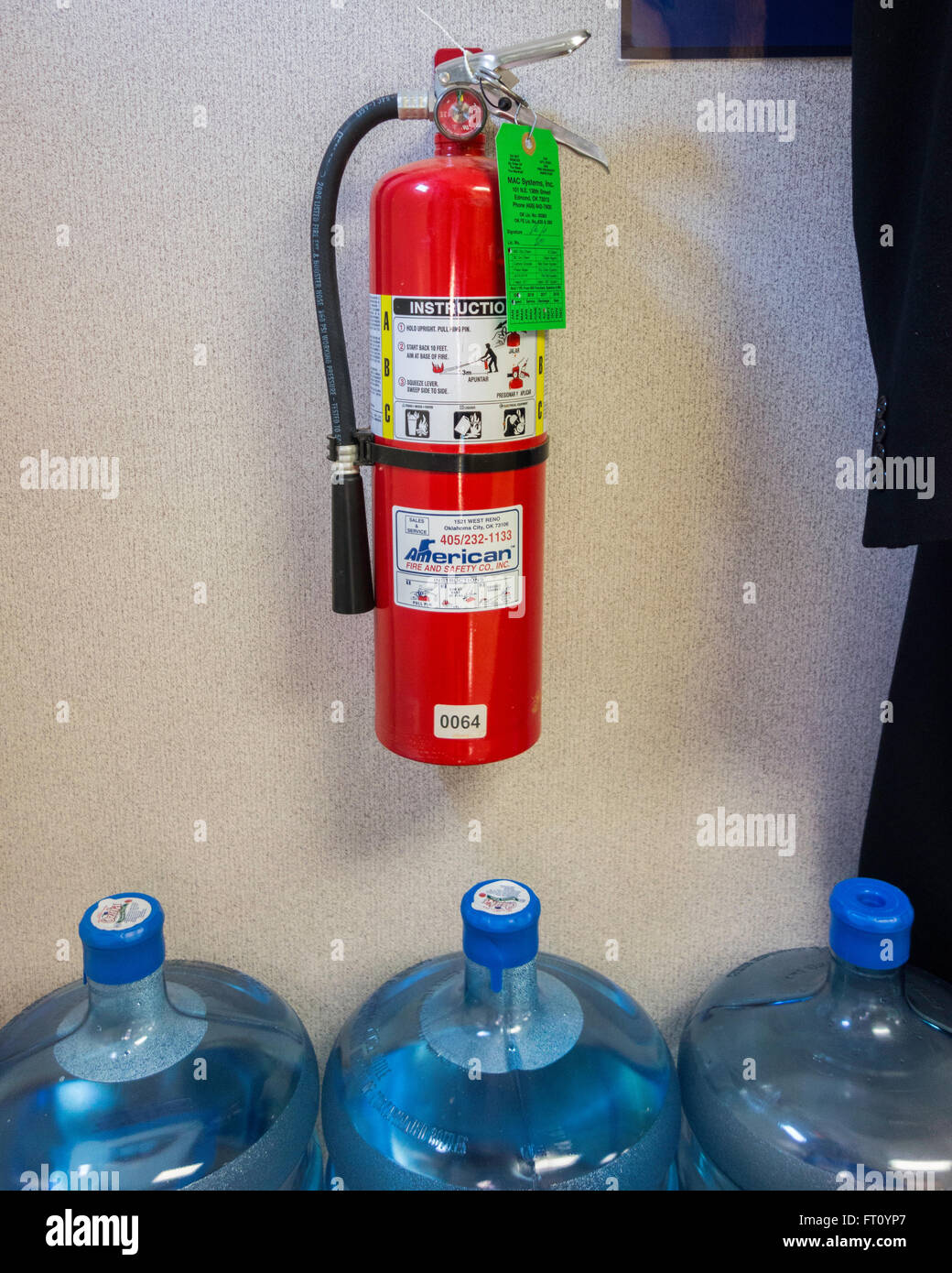 A mandatory fire extinguisher hangs on a wall over 3 five gallon water bottles in a storage area of a business. Oklahoma, USA. Stock Photo