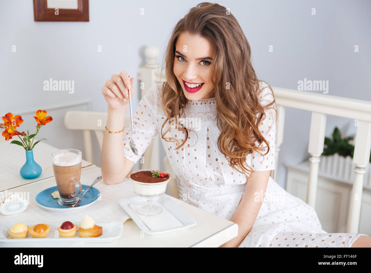 Cheerful pretty young woman with beautiful curly hair eating dessert and drinking latte in cafe Stock Photo