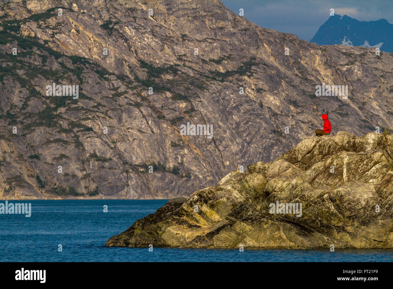 Person in a red jacket sitting on the rocky coast and enjoying the view in Glacier Bay National Park, Alaska Stock Photo