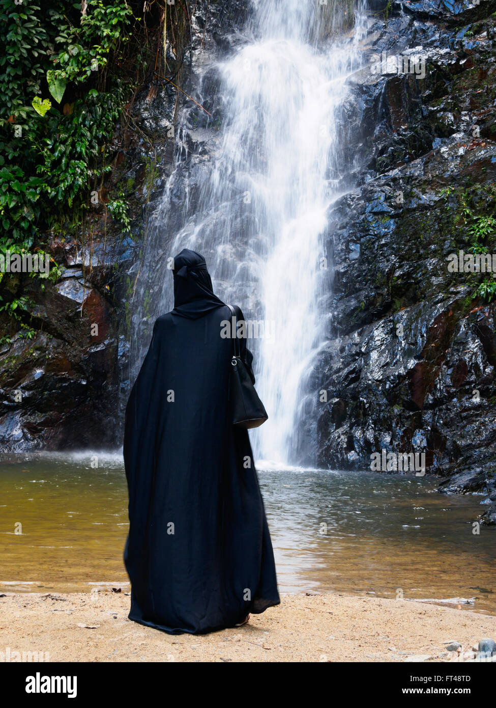 Muslim woman in full black abaya and head covering standing looking at waterfall, Langkawi Island, Kedah, Malaysi Stock Photo
