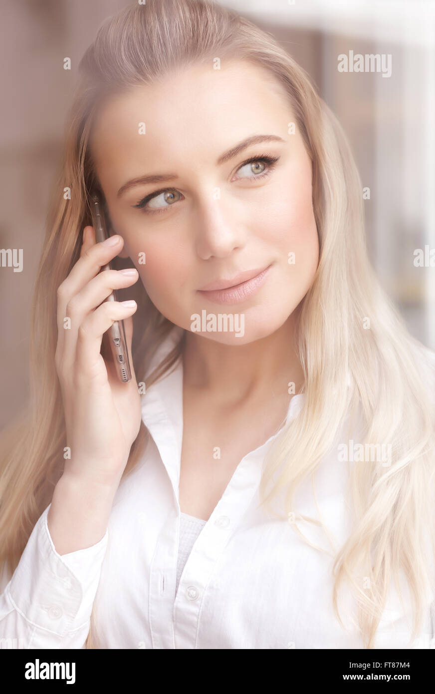 Portrait of cute blond woman standing near window at the office and talking on the phone, mobile connection Stock Photo
