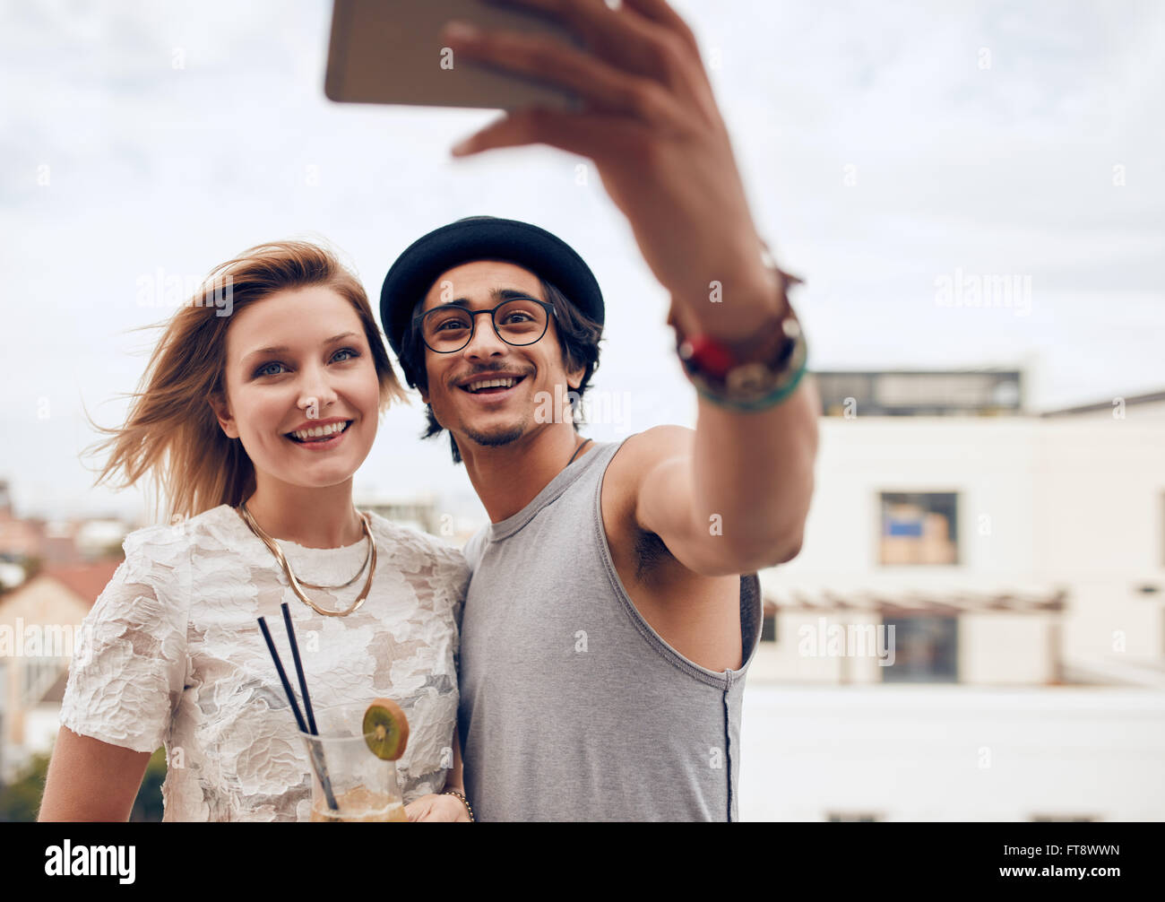 Two young friends taking self portrait with smart phone during a party. Young man and woman taking selfie at rooftop party. Stock Photo
