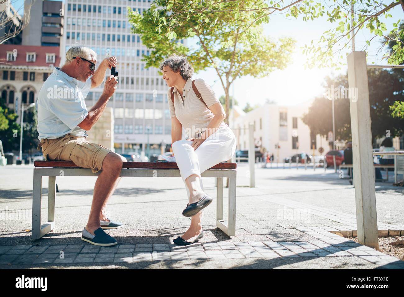 Side view of senior couple taking pictures while on vacation. Mature couple sitting outdoors on a bench photographing each other Stock Photo