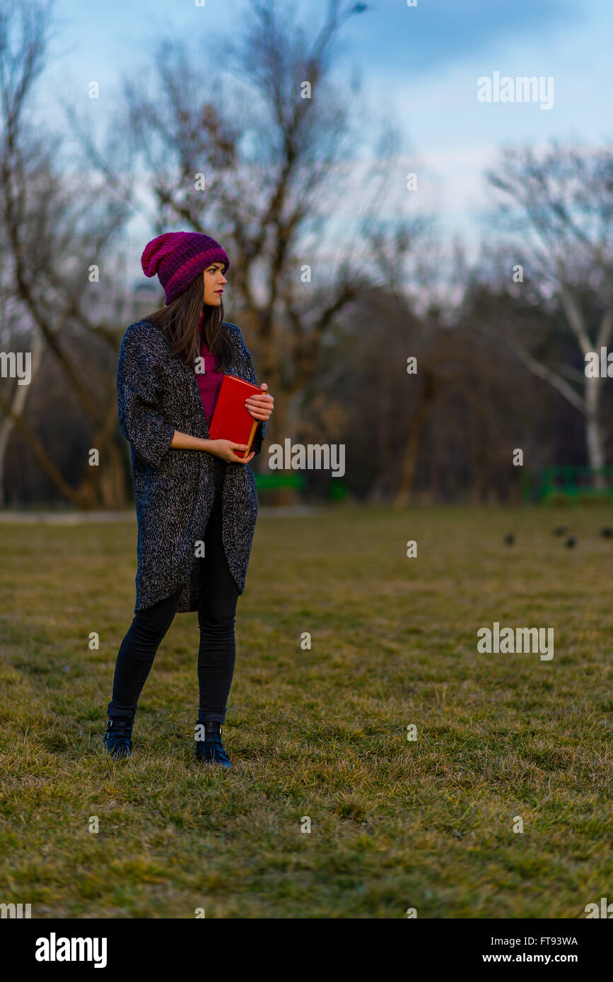 A teenager girl in purple hat holding book with red cover while standing on a lawn in a park and looking away. Stock Photo