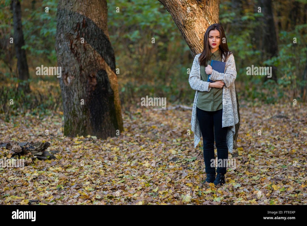 Scared by reading fairytales. A teenager girl is holding a book in an evening forest with two oak trees in the background. Stock Photo