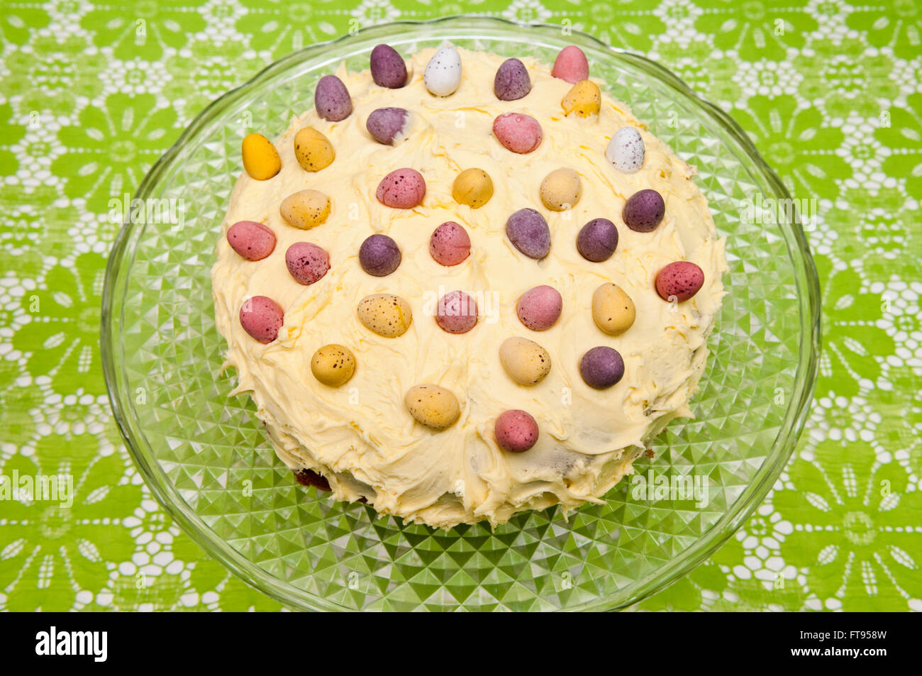 A homemade round Easter cake decorated with lemon butter cream and chocolate mini eggs on a vintage glass cake stand Stock Photo