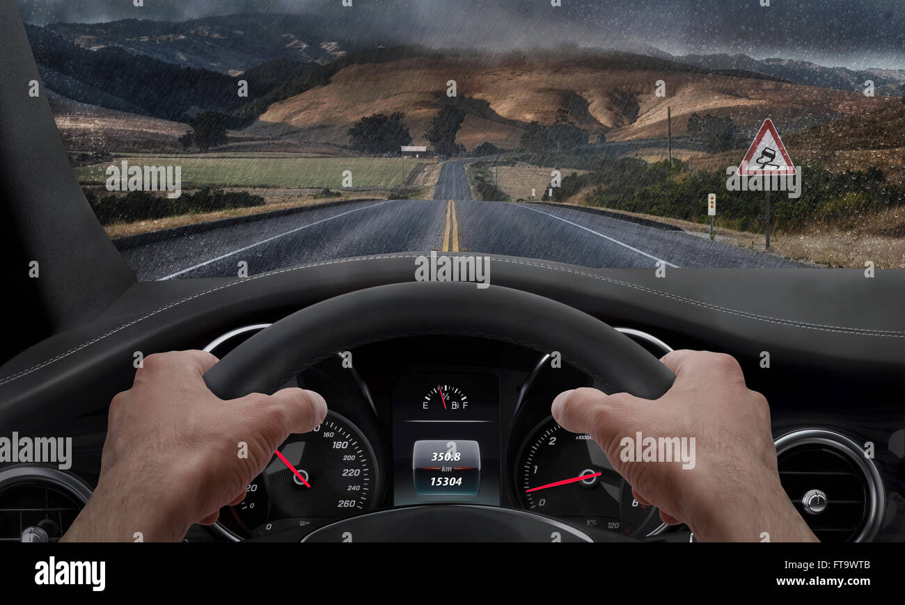 Driving in rainy weather. View from the driver angle while hands on the wheel. Alongside the road is a sign for slippery road. R Stock Photo