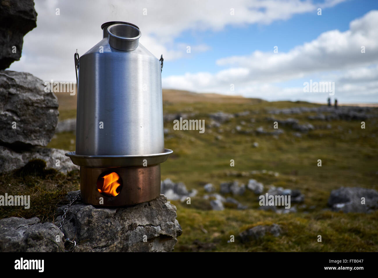 A kettle stove burning showing flames in the fire box sat on a rock with rock and grass in the background. Stock Photo