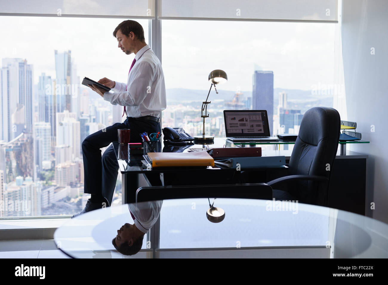Adult businessman sitting on desk in modern office and reading news on tablet pc. The man works in a skyscraper with a view of t Stock Photo