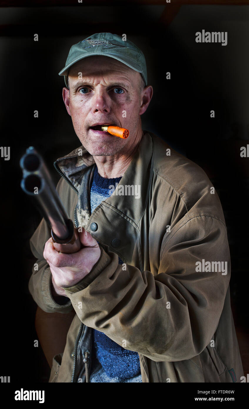 A man pointing a shotgun with a spare cartridge in his mouth Stock Photo