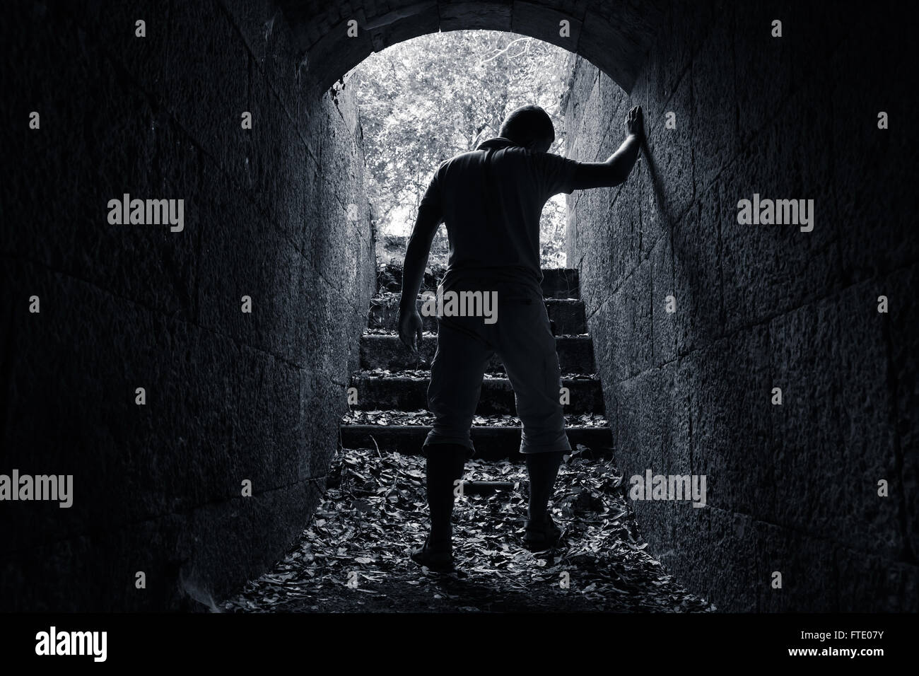Young tired man leaves dark stone tunnel with glowing end, dark blue monochrome photo Stock Photo