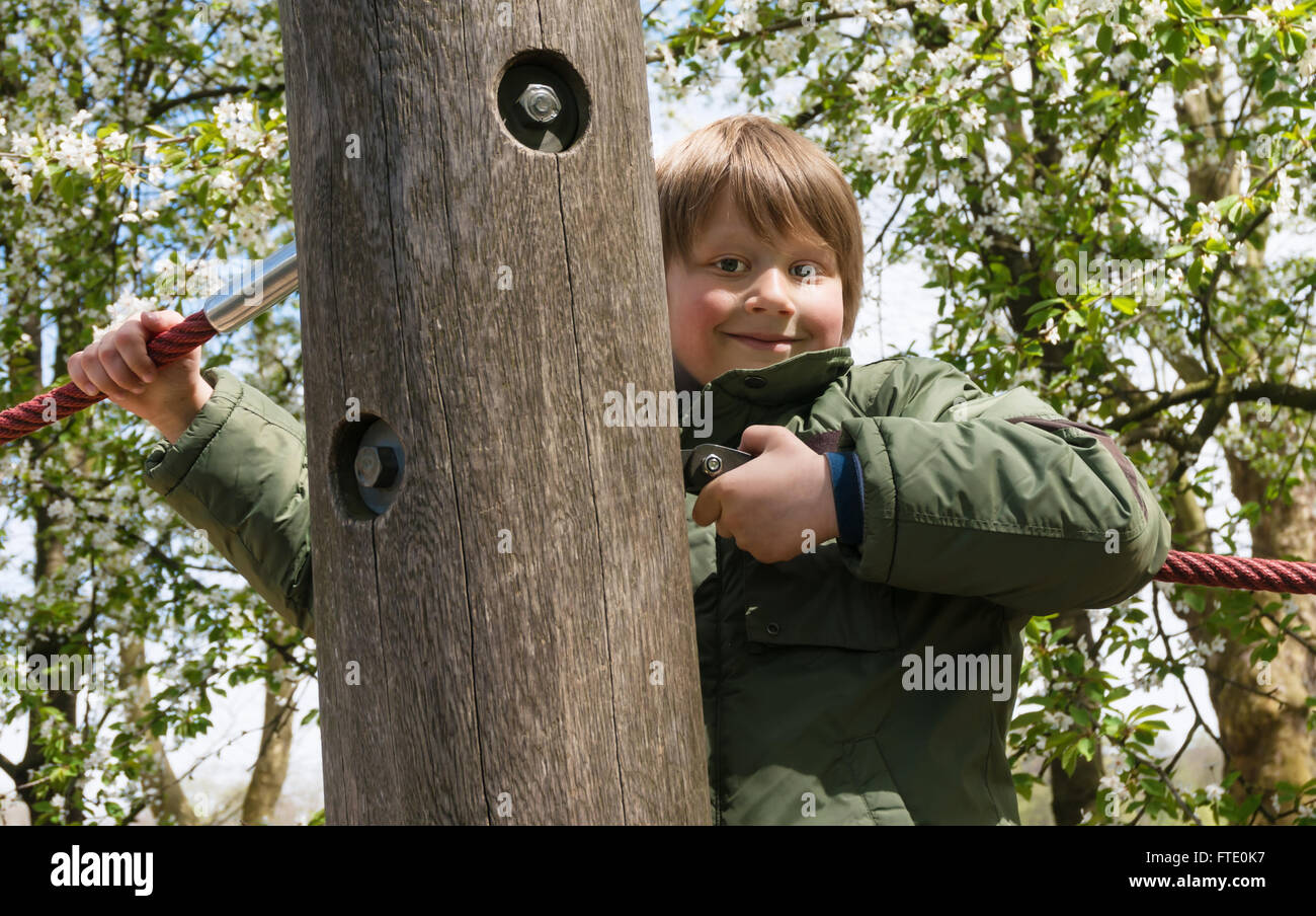 Playful blond boy peeking around a wooden post at outdoor playground Stock Photo