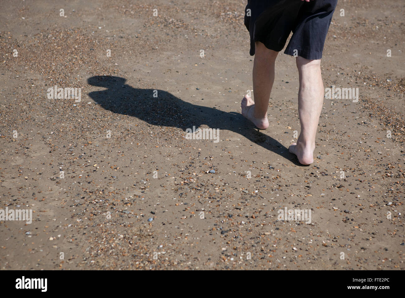 The llegs of a male walking on a beach and wading or paddling in the sea, the body throws a shadow of the figure across the sand as the person enjoys a cool walk on this hot summers day on holiday or vacation. Stock Photo