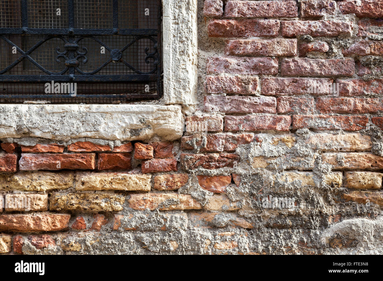 Richly coloured crumbling, exposed and decaying brickwork around a stone framed ornately barred window in a building in Venice Stock Photo