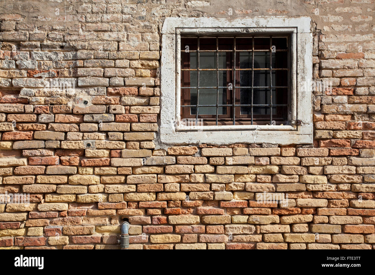 A sunlit barred stone-framed window in a wall of warm coloured crumbling brickwork on Fondamenta Nani, Venice, Italy Stock Photo