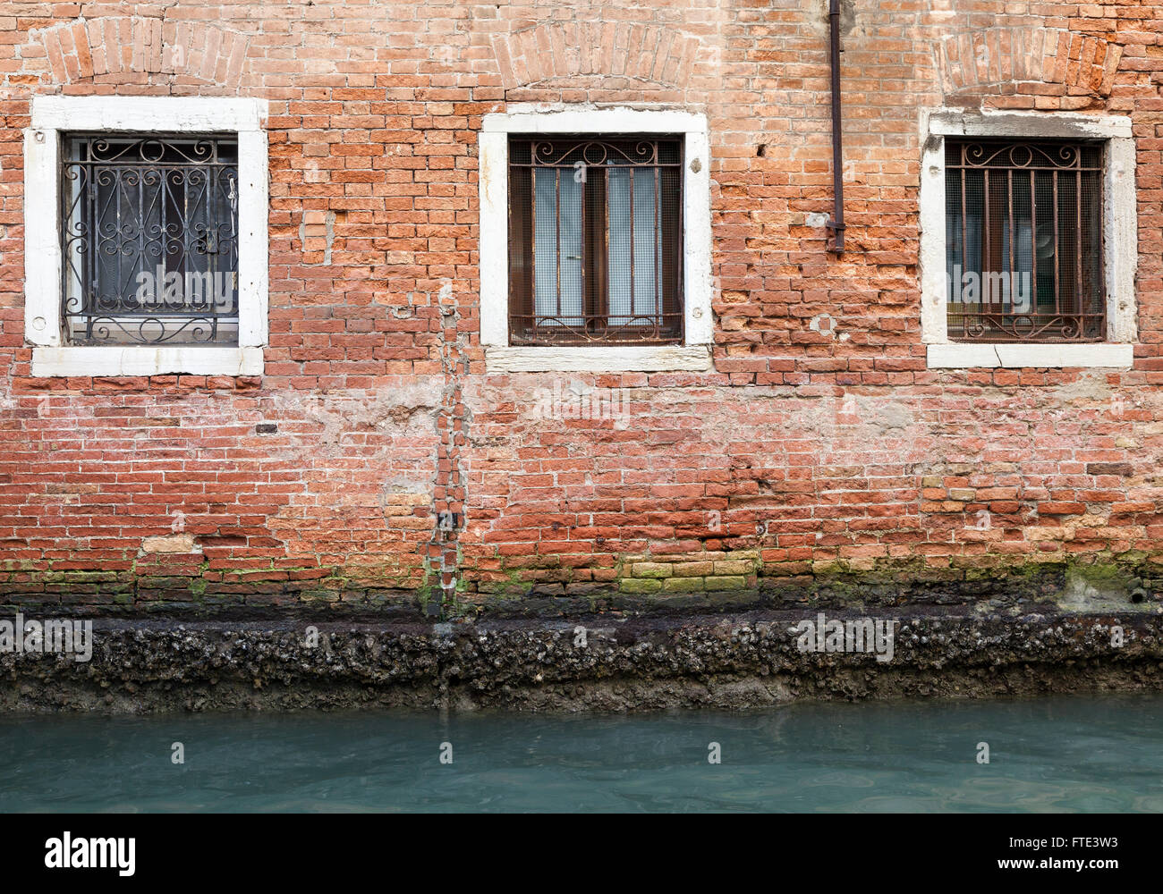 Three wrought iron barred stone-framed windows set in crumbling brickwork, green with algae, in the canal side wall of a building in Venice, Italy Stock Photo