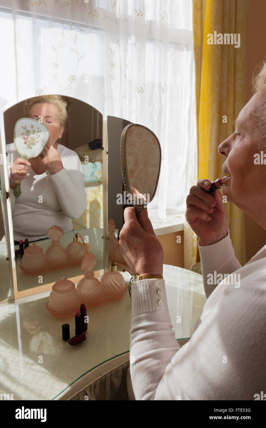 Elderly lady sat at an old fashioned dressing table applying lipstick. Stock Photo