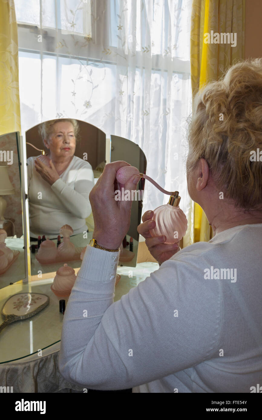 Elderly lady sat at an old fashioned dressing table applying perfume. Stock Photo