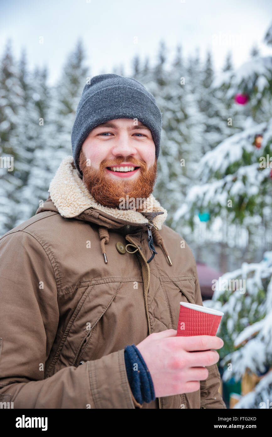 Happy young bearded man drinking hot coffee near decorated christmas tree in winter forest Stock Photo