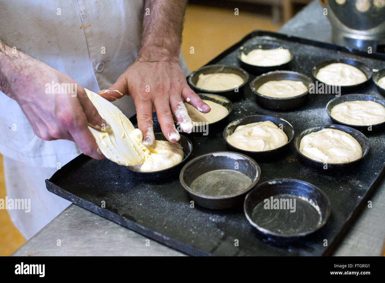 Baker making pastries in a bakery filling metal oven tins on a tray for baking with raw dough , close up of his hands Stock Photo