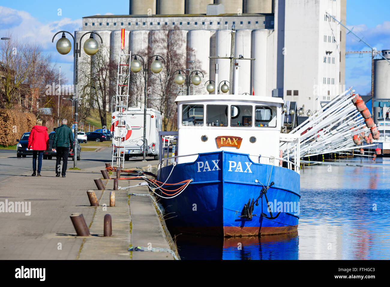 Ahus, Sweden - March 20, 2016: People walking beside a fishing boat in the marina with some industrial buildings in the backgrou Stock Photo
