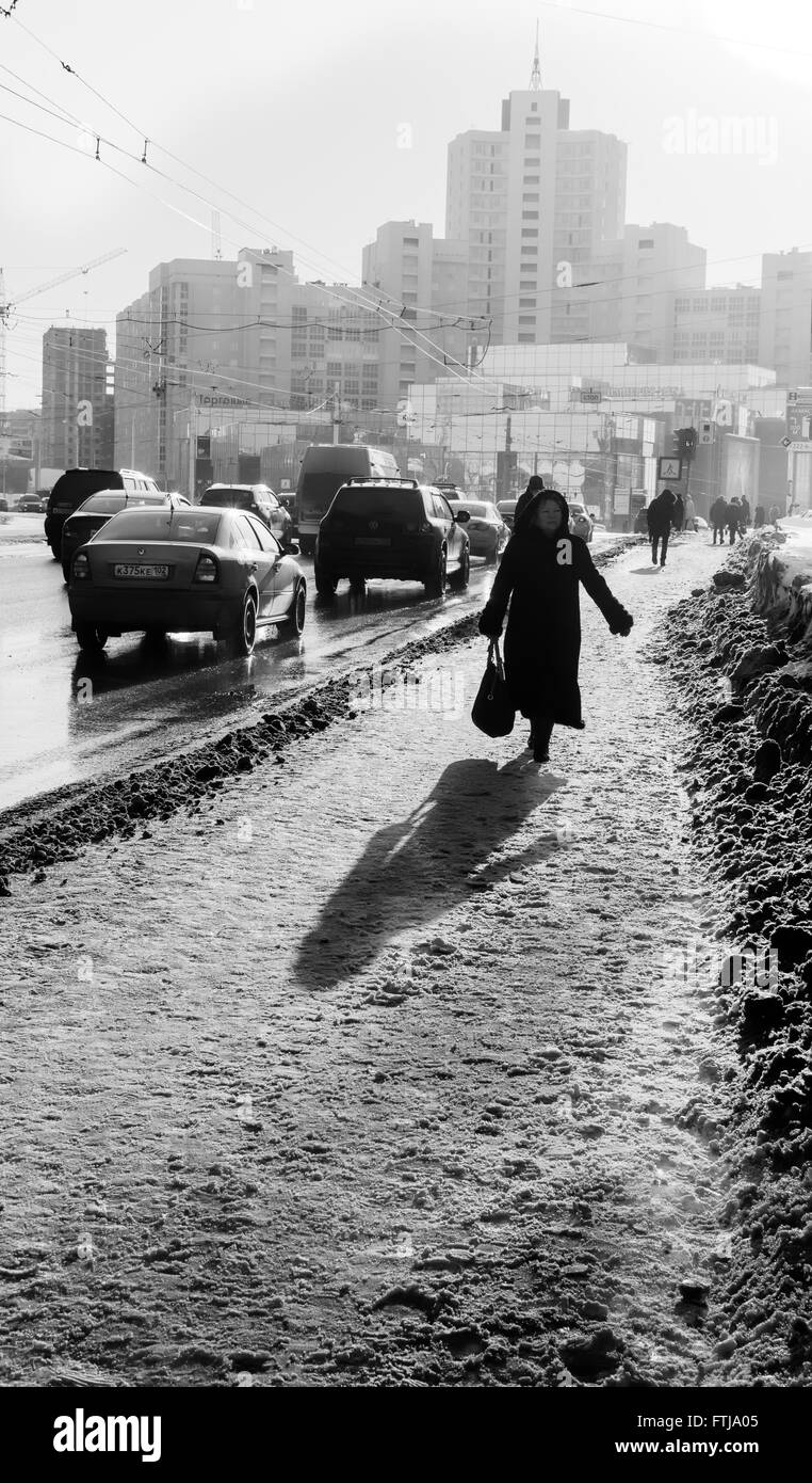 Black and white image of an old woman with shopping bags walking on a busy snow covered foot path in a city as traffic and cars Stock Photo