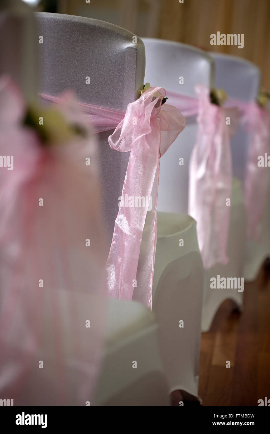White covered wedding chair backs with pink ribbon sashes and bows with wall of lights in background. Stock Photo
