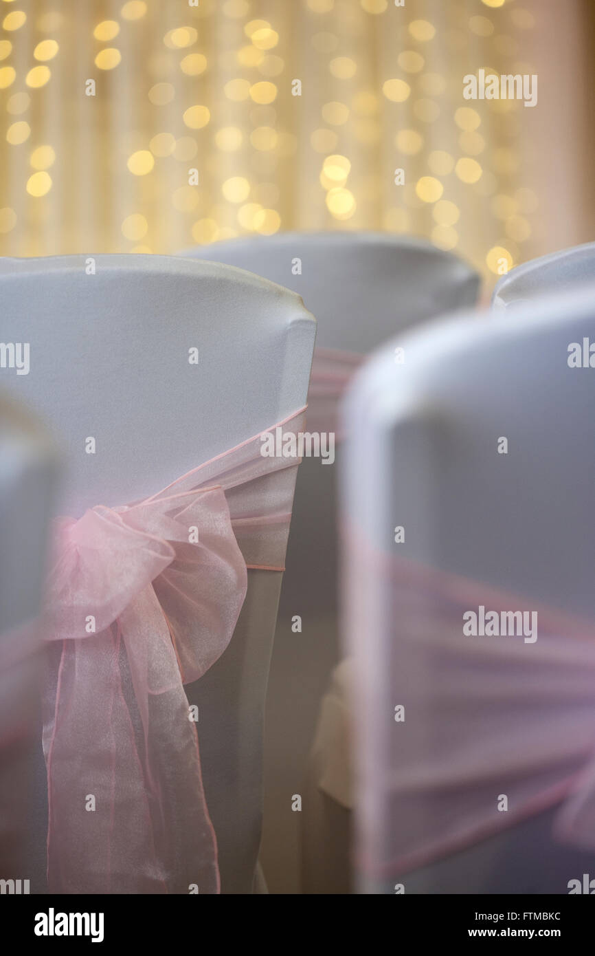 White covered wedding chair backs with pink ribbon sashes and bows with wall of lights in background. Stock Photo