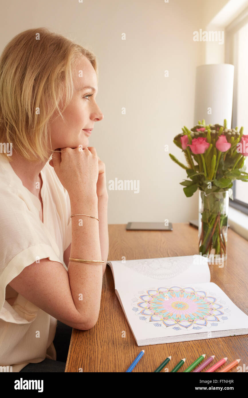 Side view of thoughtful mature woman sitting at a table with adult coloring book and pencils colors. Stock Photo