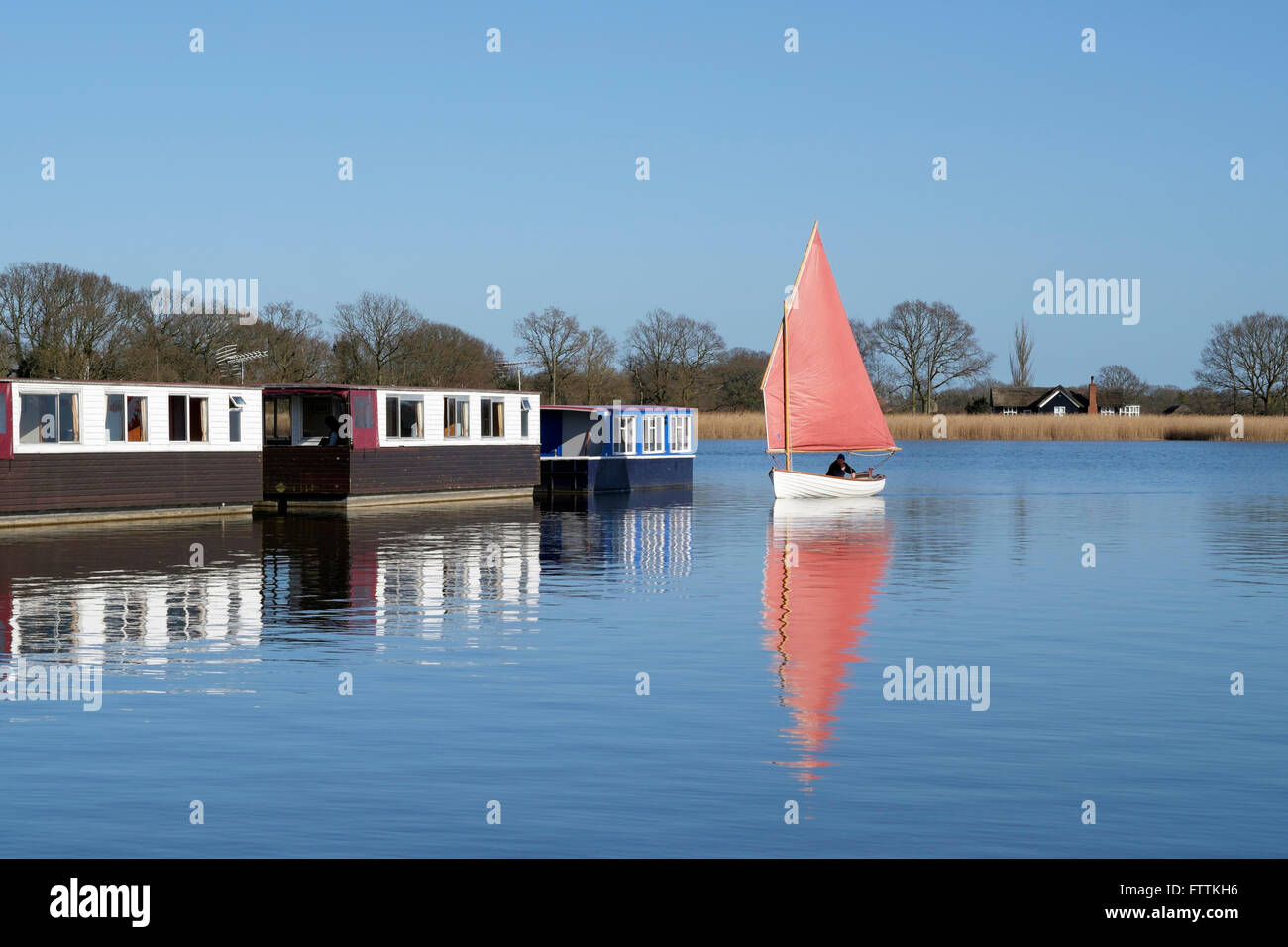 A traditional clinker hulled sailing dinghy sails past a row of houseboats on Hickling Broad, Norfolk, England, UK Stock Photo