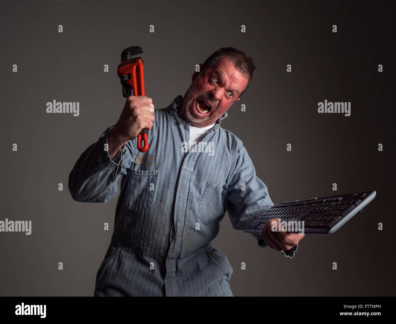 Photograph of an angry repairman hollering and about to smash a computer keyboard with an orange pipe wrench (humor). Stock Photo