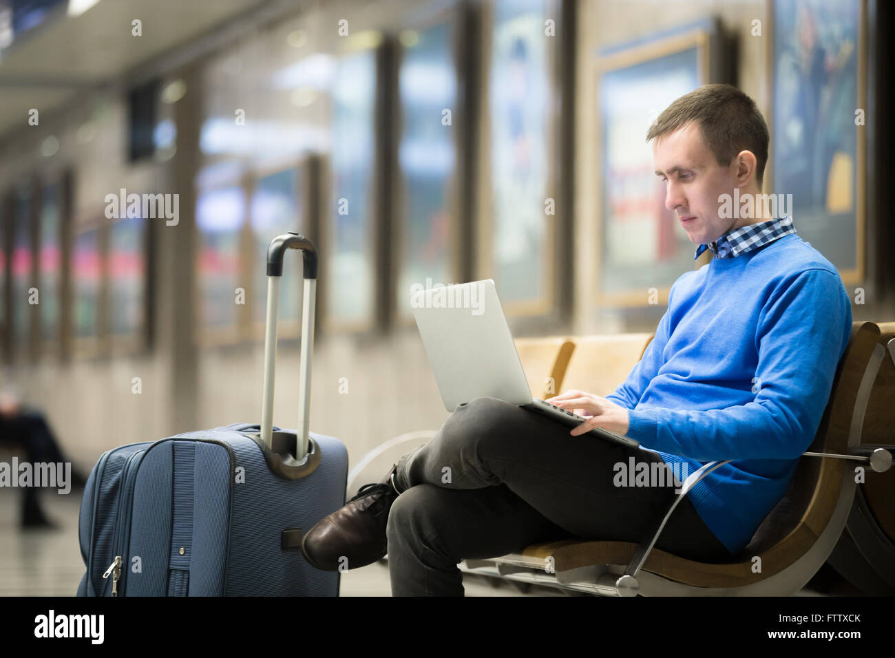 Portrait of young handsome man wearing casual style clothes sitting on the bench in modern airport using laptop. Passenger Stock Photo