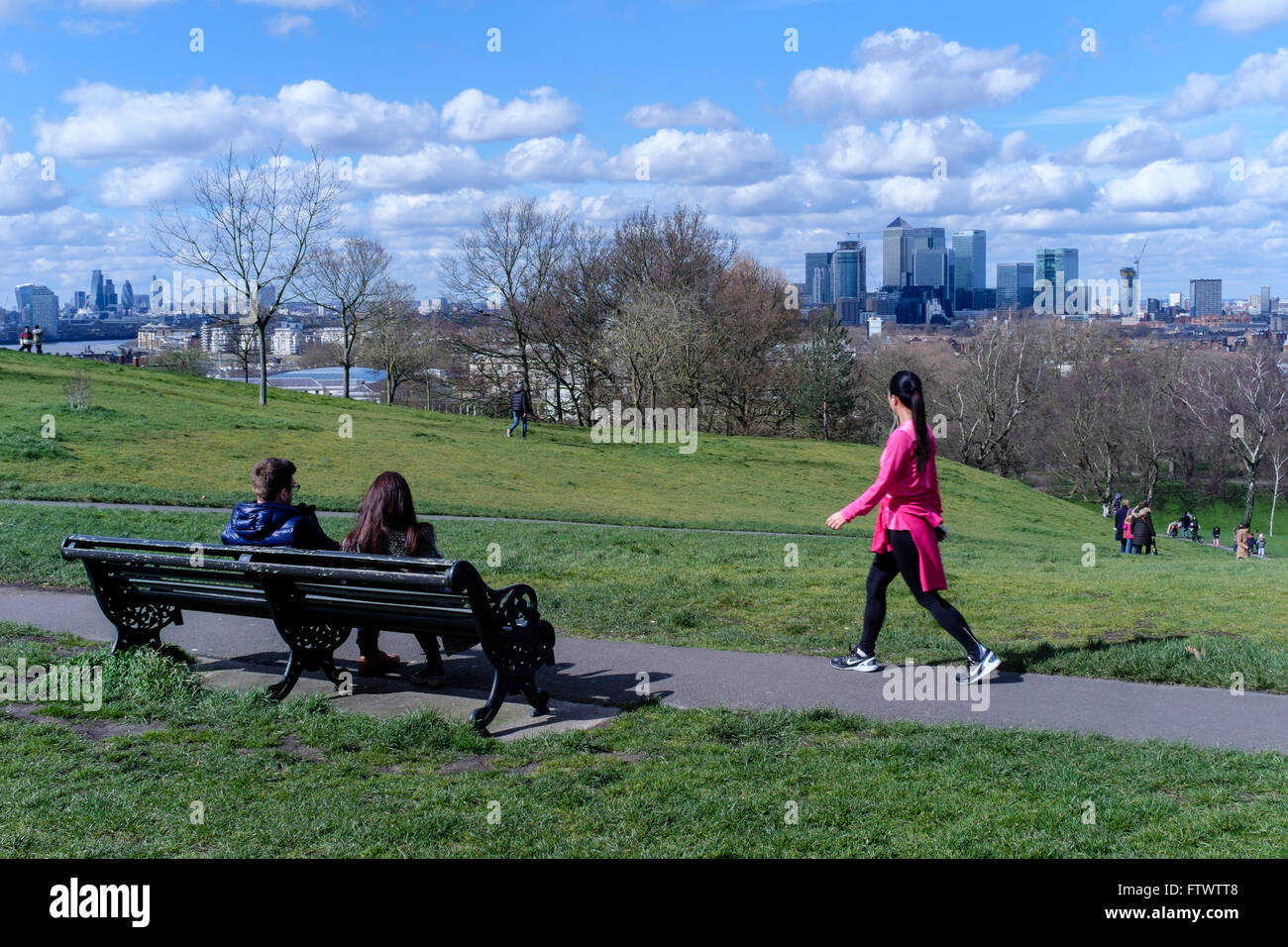 Young woman in exercise clothing walks past couple sitting on bench, Greenwich Park, London, UK Stock Photo