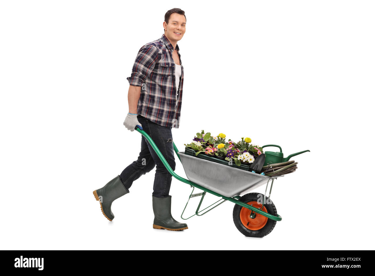 Young gardener pushing a wheelbarrow full of gardening equipment and flowers isolated on white background Stock Photo