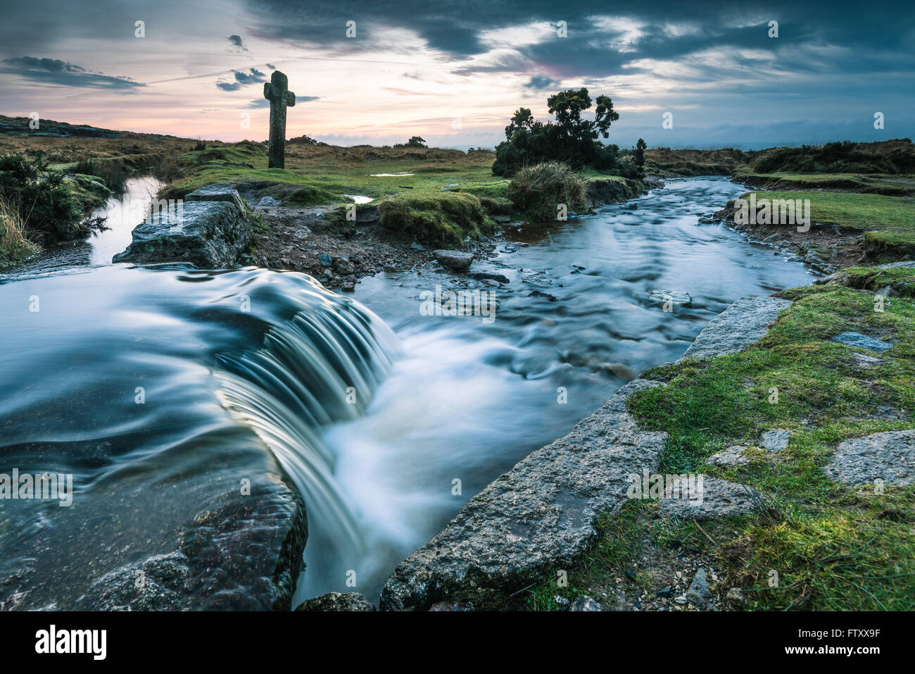 Water flowing in wild creek, ancient Cross in background in Dartmoor Park, UK Stock Photo