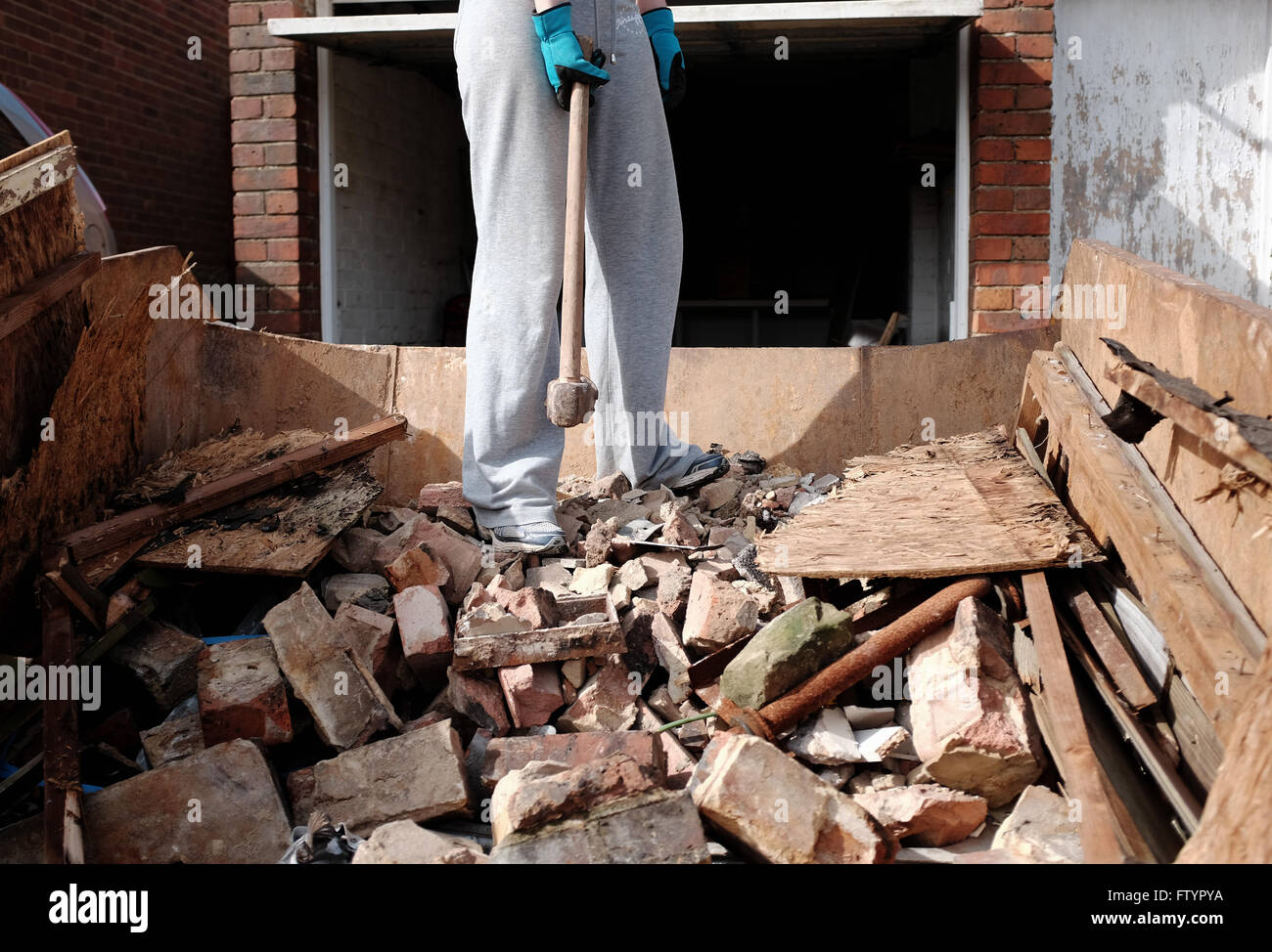 Young woman in her 20s doing DIY at her bungalow home with a sledgehammer in a skip Stock Photo