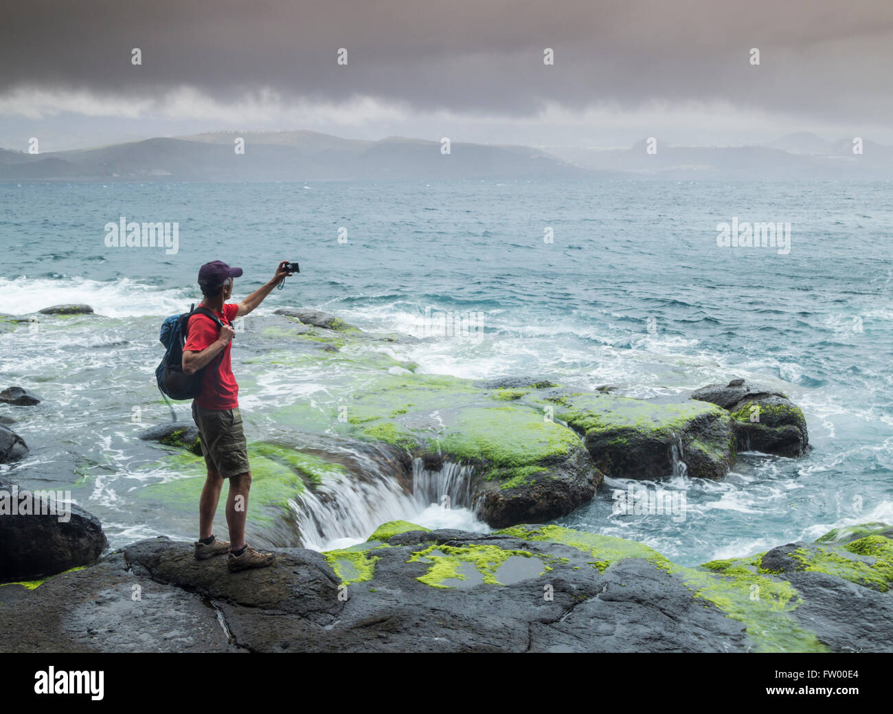 Las Palmas, Gran Canaria, Canary Islands, Spain, 30th March 2016. Weather: Warnings have been issued for heavy rain and rough seas as a mass of cold air sweeps across the Canary Islands. PICTURED: A hiker on the rugged north coast of Gran Canaria takes a selfie as Atlantic waves crash on the rocks and storm clouds pass over the mountains in the distance. Credit:  Alan Dawson News/Alamy Live News Stock Photo