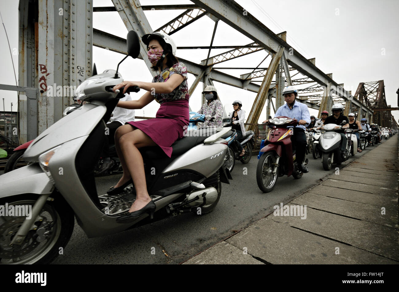 Woman wearing a face mask and a skirt riding a scooter during rush hour on Long Bien bridge in Hanoi, Vietnam Stock Photo