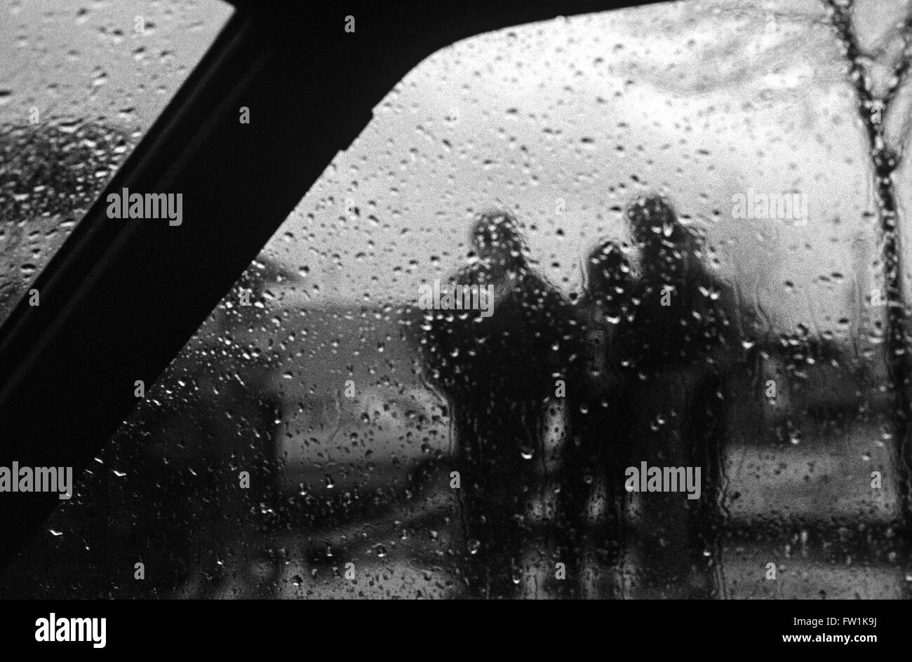 Three people waiting on street to cross road in rain blurred by interior car window Stock Photo