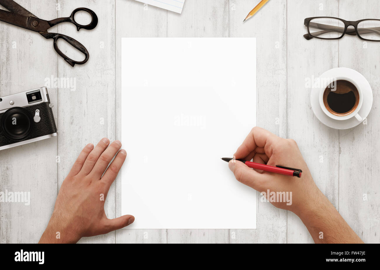 Man write with red pen on isolated, white paper. Coffee, glasses, camera, notepad, pencil on wooden table. Top view. Stock Photo