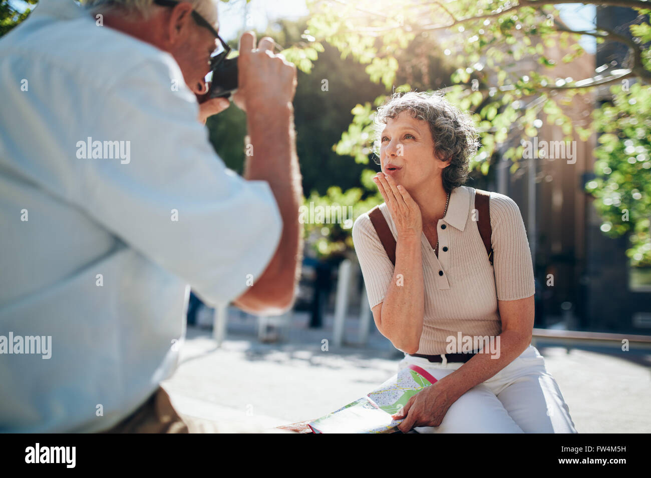 Beautiful senior woman blowing a kiss to camera while sitting outdoors on a bench in the city. Woman being photographed by his h Stock Photo
