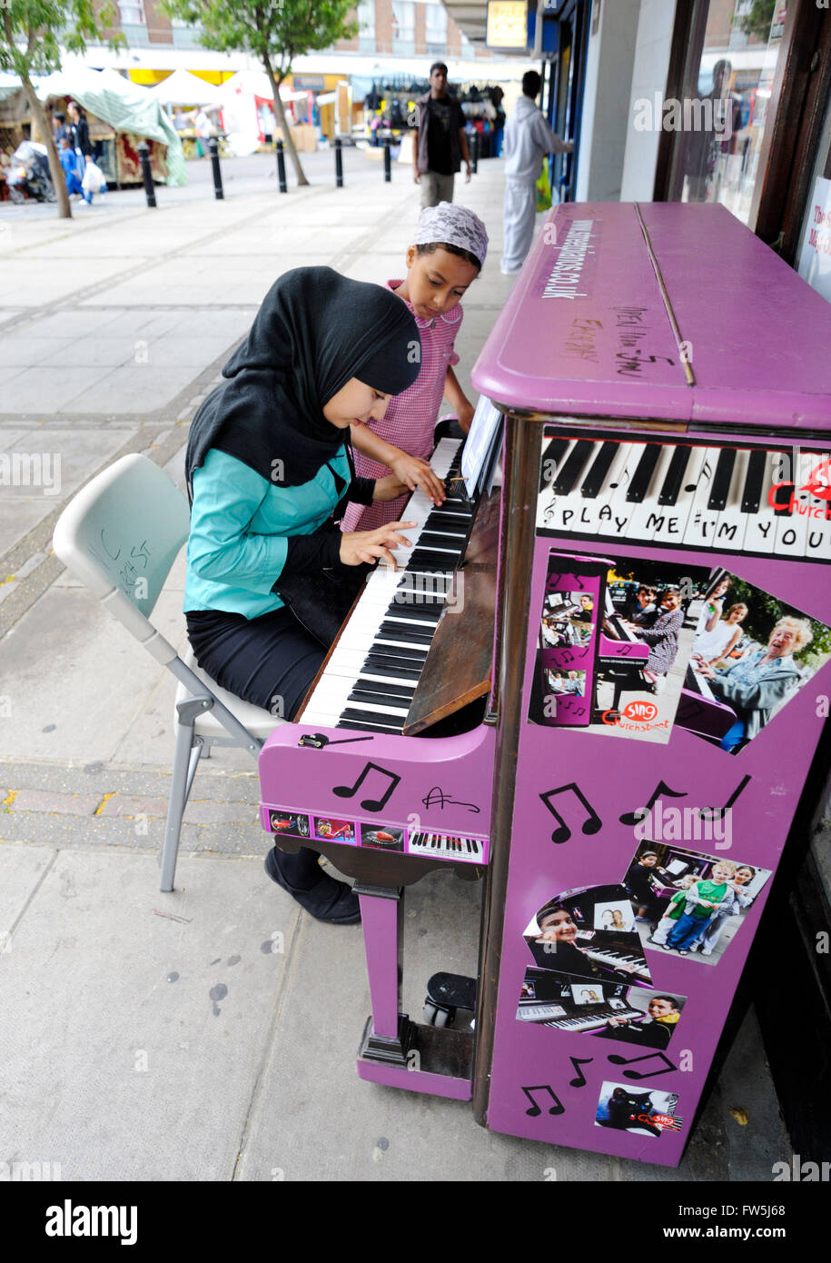 Asian girls on street market playing old upright piano (by Chappell), painted purple. Part of Streetpiano.co.uk 'Play me, I'm yours' scheme by artist Luke Jerram, for Sing London and the City of London Festival. 30 pianos are left in public areas arond London, summer 2009 Stock Photo