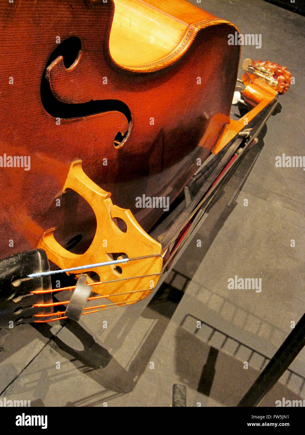 5-string double bass resting on its side, with bridge prominent, mute in 'ready' position below bridge, bow tucked away under strings, and shadows on floor, including shadow of music stand. Stock Photo