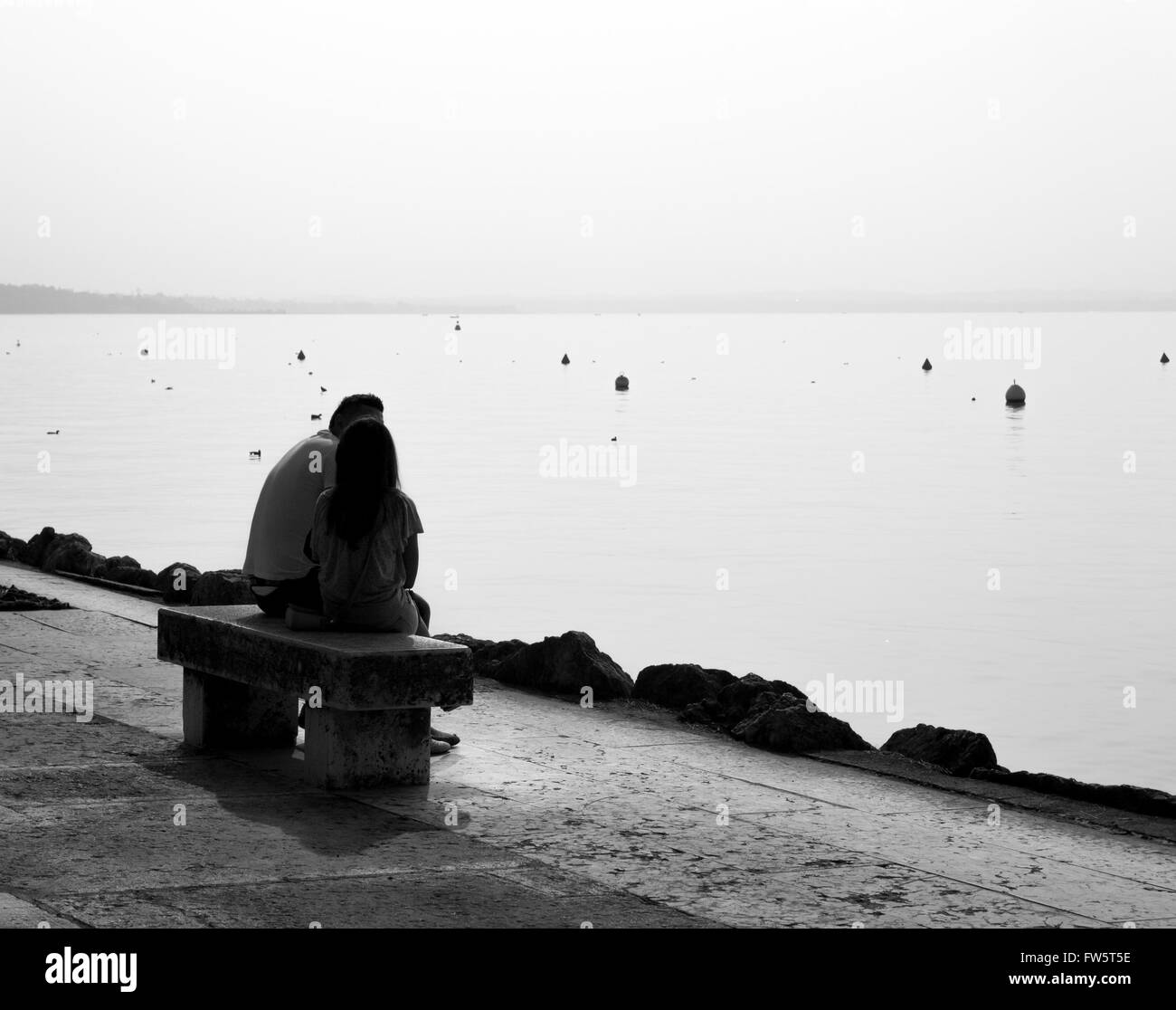 Lovers seated on a bench on the lake shore backlit. Stock Photo