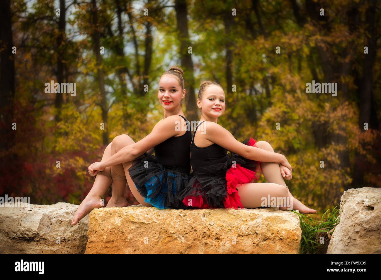 Two Sisters Pose back to back in ballet tutus on a rock in the park in Autumn Stock Photo